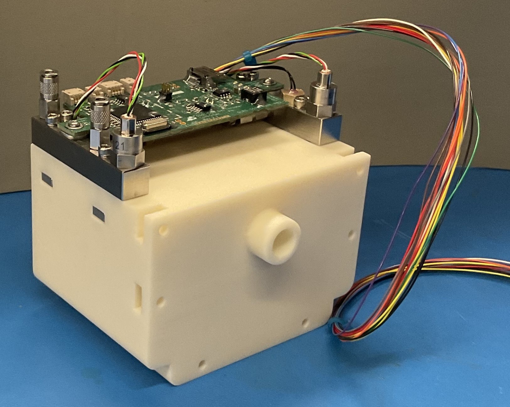 Isometric view of a tan-colored cube with a nozzle facing out of one side; PCB, wires, and sensors mounted to the top with metal brackets